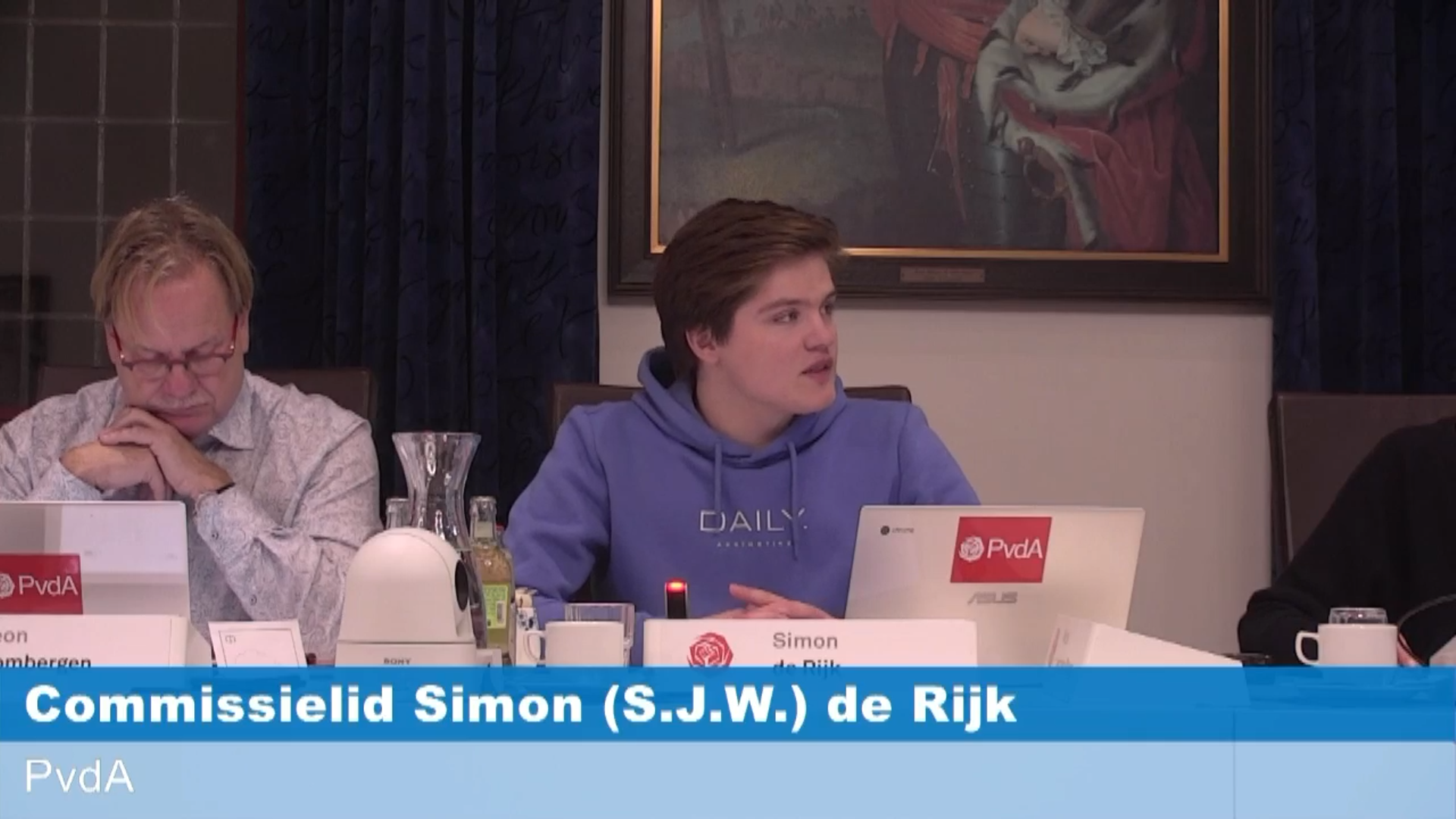 Commissielid Simon de Rijk zit met een chromebook voor zich, pratend, aan de raadstafel en kijkt naar rechts.