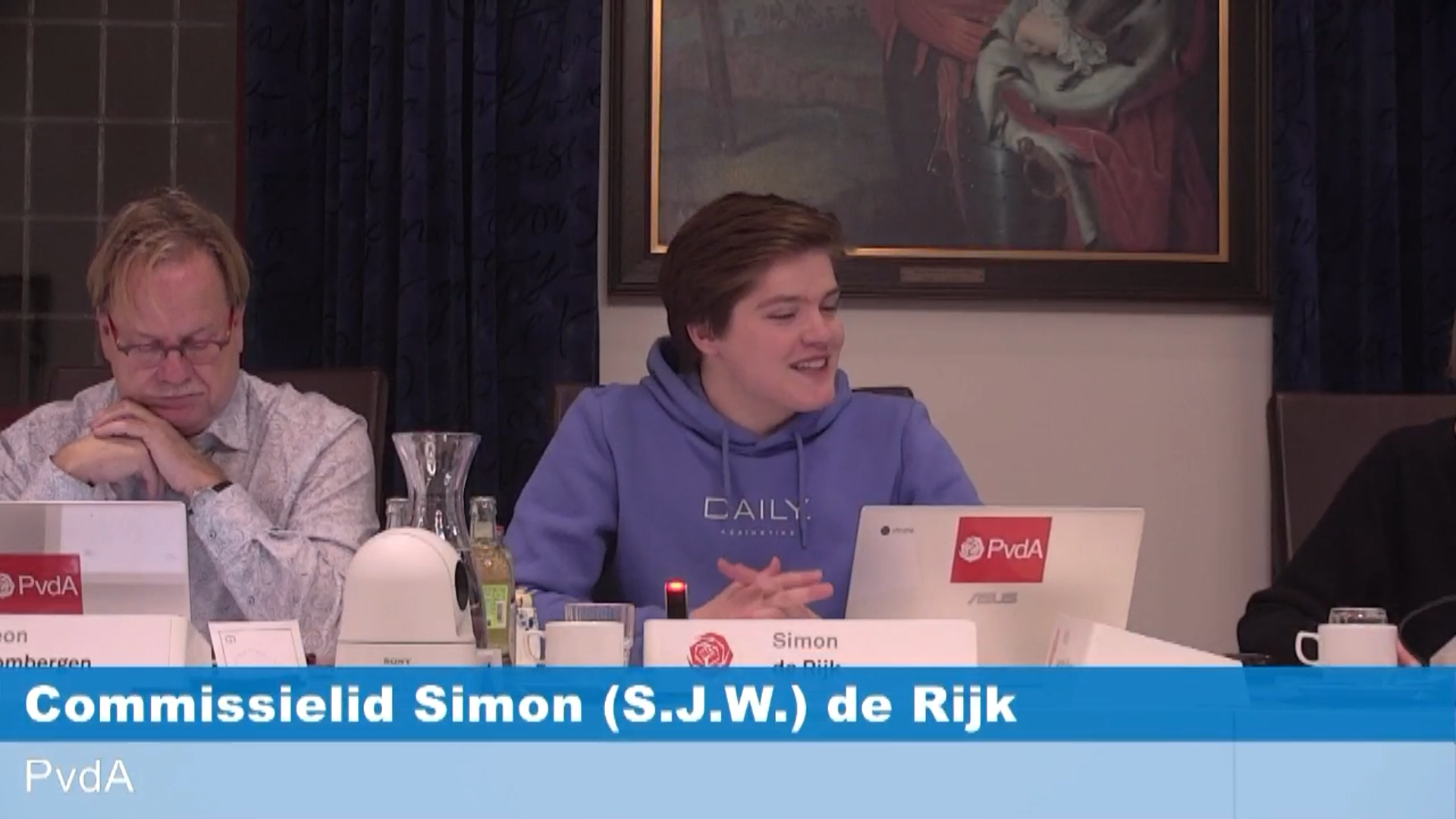 Commissielid Simon de Rijk zit met een chromebook aan de raadstafel, glimlachend, pratend en met zijn handen in elkaar. Achter hem is een schilderij te zien.
